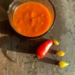 En glasskål fyldt med stærk, orange chili sauce med habanero. Ved siden af skålen ligger en rød chili og tre små gule habanero-chilier på en rustik overflade i stærkt sollys.