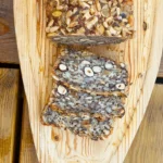 Et mørkt kernebrød med hasselnødder skåret i skiver og anrettet på et lyst træbræt. Man ser tydeligt de hele nødder og mange kerner.