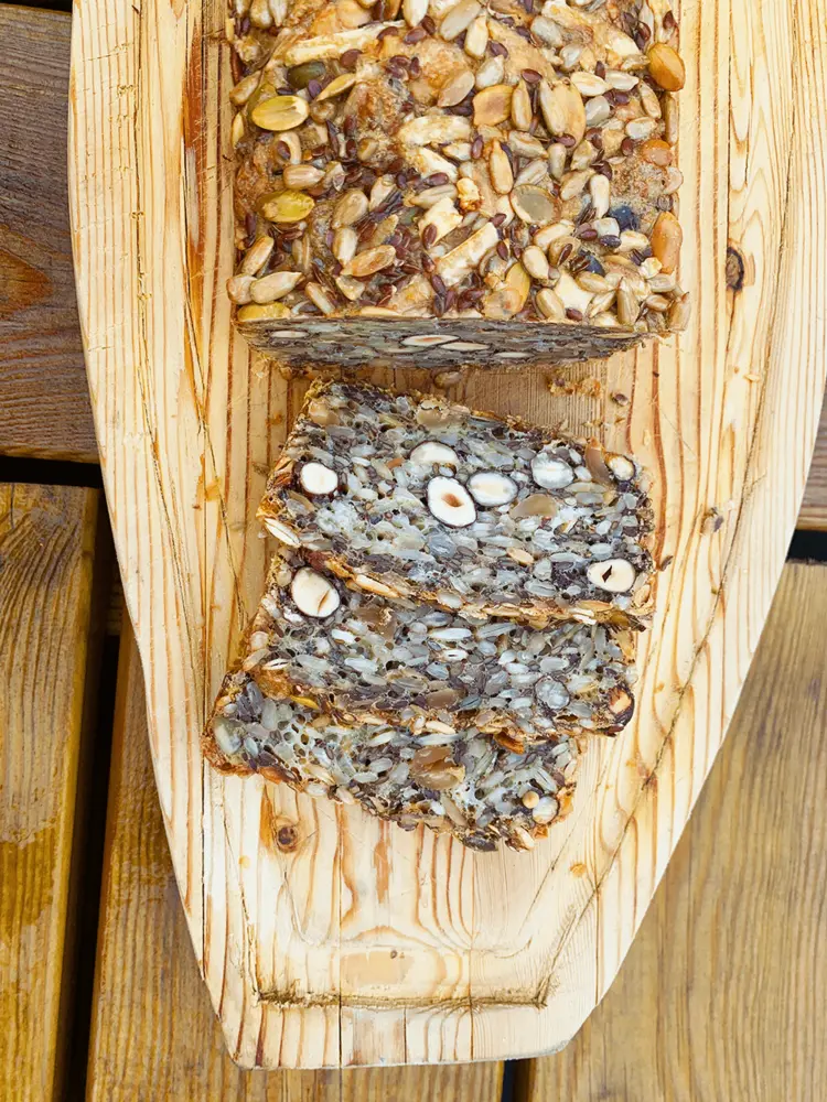 Et mørkt kernebrød med hasselnødder skåret i skiver og anrettet på et lyst træbræt. Man ser tydeligt de hele nødder og mange kerner.