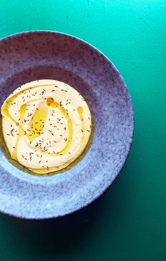 Hummus med olivenolie i blå skål på grøn baggrund - skålen er skåret lidt