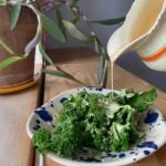 En hvid kande hælder cremet vinaigrette dressing over en tallerken med frisk grønkålssalat.