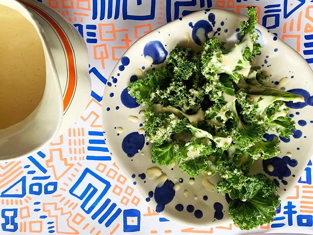 Set fra oven: En cremet vinaigrette dressing hældes fra en kande over frisk grønkål på en hvid tallerken med blå pletter. Tallerkenen står på en dug med blåt og orange mønster.