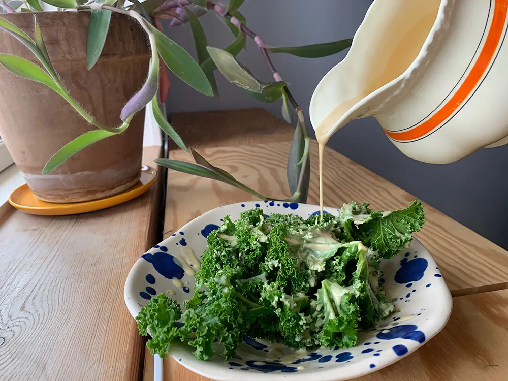 En hvid kande hælder cremet vinaigrette dressing over en tallerken med frisk grønkålssalat.